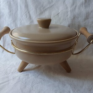 May include: A beige ceramic serving dish with a lid and wooden handles. The dish sits on a gold metal stand with three wooden legs.