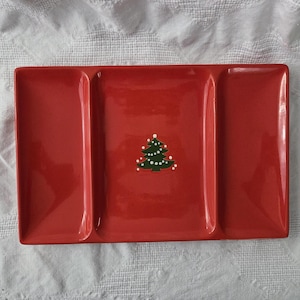 May include: A rectangular red ceramic serving tray with three distinct sections. The center section features a small, dark green Christmas tree graphic decorated with white dots representing lights or snow, centered on the glossy red surface.