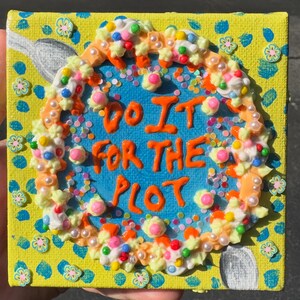 May include: A square yellow canvas featuring a blue center circle with the orange text DO IT FOR THE PLOT. The design is bordered by textured white and orange icing-like piping, colorful sprinkles, small pearls, and tiny flower embellishments.