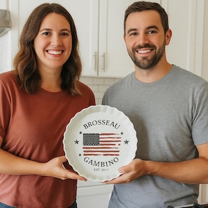 May include: A white ceramic fluted pie dish featuring a graphic of the American flag in the center. The text BROSSEAU is printed above the flag, GAMBINO is printed below, and EST 2017 is at the bottom. Four black stars surround the flag design.
