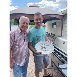 May include: Two people stand outside, one holding a white oval plate with the text "Grill Master Manning" and a grill utensil graphic. The person holding the plate is wearing a blue and white striped shirt and khaki shorts. The other person is wearing a blue shirt with a graphic and a striped shirt.