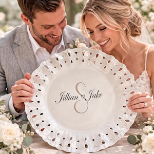 May include: A white ceramic plate with a decorative, openwork edge. The plate features the names "Jillian & Jake" in elegant script. The plate is held by a couple, suggesting a wedding or special occasion.
