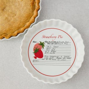 May include: A white ceramic fluted pie dish featuring a red strawberry illustration and a printed recipe for Strawberry Pie crust. The text includes ingredients like flour, oil, salt, sugar, and milk, with baking instructions for 8-10 minutes at 400 degrees.