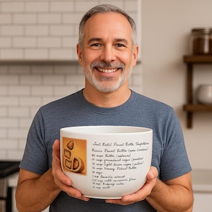 May include: A white ceramic mixing bowl with a recipe for "Aunt Ballie's Peanut Butter Temptation" printed on the side. The bowl features a graphic of peanut butter cookies and is held by a person.