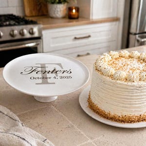 May include: A white ceramic cake stand with the name "Fenters" and the date "October 4, 2025" printed in black. A frosted cake with a crumb crust sits on a white plate. The cake has white frosting and decorative swirls.