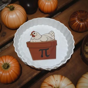 May include: A white ceramic fluted pie dish featuring a central graphic of a white chicken sitting inside a brown terracotta pot. The pot is decorated with a black mathematical pi symbol. The dish has a scalloped edge and a smooth, glossy finish.