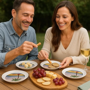 May include: A wooden table set for a snack with small white plates featuring a sunset and palm tree design. The table holds cheese, crackers, and grapes. A man and woman are seated at the table, enjoying the food and drink.