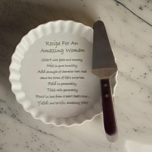 May include: A white ceramic fluted tart dish featuring the printed text Recipe For An Amazing Woman, Start with faith and honesty, Mix in pure humility, Add strength of character, Fold in personality, TOSS with generosity, Pour in love, Yield: one terrific, amazing YOU, paired with a metal cake server with a dark brown handle.