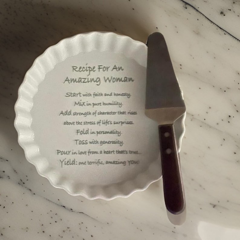 May include: A white ceramic fluted tart dish featuring the printed text Recipe For An Amazing Woman, Start with faith and honesty, Mix in pure humility, Add strength of character, Fold in personality, TOSS with generosity, Pour in love, Yield: one terrific, amazing YOU, paired with a metal cake server with a dark brown handle.