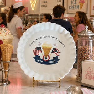 May include: A white ceramic pie dish with a scalloped edge featuring a central graphic of an ice cream cone, two flags, and the text Our Ice Cream Social 50th Anniversary 1976 2026. The dish is displayed on a gold wire stand on a marble counter.