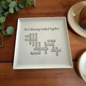 May include: A square, off-white ceramic plate with the text "Bev's Blessings Linked Together" and a crossword puzzle design featuring names. The names are in black, and the plate has a slightly raised edge. The plate is on a wooden surface.
