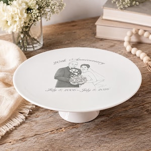 May include: A white ceramic cake stand featuring a central gray illustration of a couple in wedding attire holding a bouquet. The text reads 20th Anniversary and July 8, 2006 - July 8, 2026. The stand has a smooth, circular top and a pedestal base.