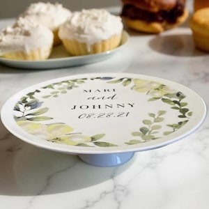 May include: White cake stand with a floral design and the text "MARI and JOHNNY 08.28.21".