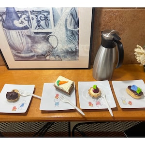 May include: Four small desserts on white plates with a child's drawing on each plate. The desserts are a chocolate cake, a carrot cake, a pastry with a red berry, and a pastry with blue frosting.