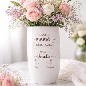 May include: A white ceramic vase filled with pink and white roses, baby's breath, and other flowers. The vase has the words "FIRST mamá Michelle Cynthia NOW abuela" and the names "River" and "Esmeralda" printed on it.