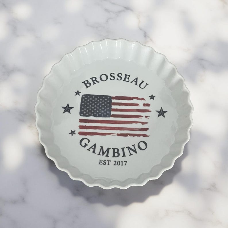 May include: A white ceramic fluted tart dish featuring a central graphic of a distressed American flag. The text BROSSEAU is printed above the flag, GAMBINO is printed below, and EST 2017 is at the bottom, all surrounded by four small stars.