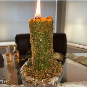 Puede incluir: Una vela verde con una capa de hierbas secas en la parte superior est&aacute; encendida y ardiendo. La vela est&aacute; sentada sobre una base cubierta de papel de aluminio plateado.