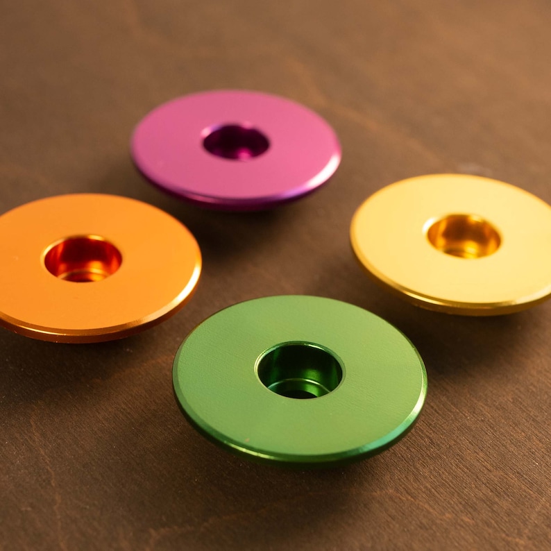May include: Four colorful, metallic bottle stoppers in shades of orange, green, purple, and gold. Each stopper has a circular design with a central hole. The stoppers are arranged on a dark brown wooden surface.