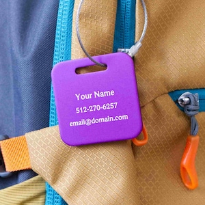 May include: A purple luggage tag with the text "Your Name", a phone number, and an email address. The tag is attached to a backpack with a metal cable. The backpack is tan and blue with orange accents.