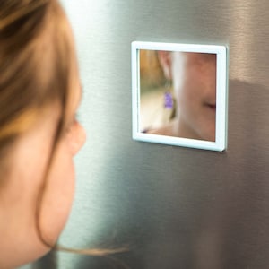 May include: A white framed mirror with a reflection of a person's face and a purple earring. The mirror is attached to a stainless steel surface.