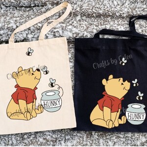 May include: Two tote bags, one cream and one black, each with a cartoon illustration of Winnie the Pooh in a red shirt, looking at bees. A honey pot with the word "HUNNY" is also shown. The text "Crafts by Colon" is printed on each bag.