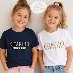 Könnte beinhalten: Ein dunkelblaues T-Shirt und ein weißes T-Shirt, jeweils mit dem Schriftzug KITAKIND in bunten Buchstaben über einer Reihe farbiger Wimpel. Die T-Shirts haben kurze Ärmel und einen Rundhalsausschnitt mit einem Grafikdruck auf der Brust.