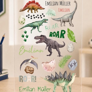 Könnte beinhalten: Eine klare Acrylplatte auf einem Holzsockel mit bunten Illustrationen verschiedener Dinosaurier, Eier, Fußabdrücke und Blätter. Der Text enthält den Namen EMILIAN MÜLLER, den Namen EMILIAN und das Wort ROAR in verschiedenen Schriftarten.