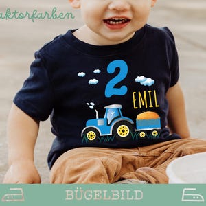 Könnte beinhalten: Ein marineblaues kurzärmeliges T-Shirt mit einem blauen Traktor, gelben Rädern und einem Anhänger. Die Zahl 2 ist blau, der Name "EMIL" gelb und weiße Wolken. Der Text "3 Traktorfarben" steht oben.