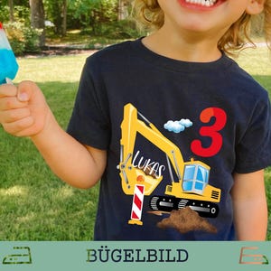 Könnte beinhalten: Marineblaues T-Shirt mit gelbem Bagger-Motiv, dem Namen "Lukas" und der Zahl "3" in Rot. Ein rot-weiß-blaues Eis am Stiel wird in der Hand gehalten. Das T-Shirt ist für ein Kind.