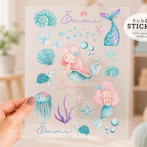Könnte beinhalten: Ein transparenter Bogen mit Rubbelstickern, der Aquarellillustrationen von zwei Meerjungfrauen mit rosa Haaren, Meeresschildkröten, Quallen, Muscheln, Seesternen und Blasen zeigt. Der Name Emmi ist an drei Stellen in Schreibschrift gedruckt.