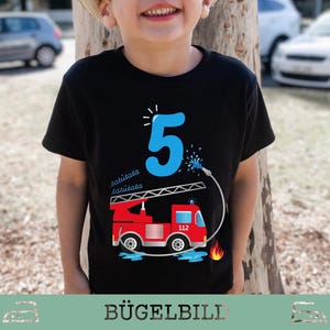 Könnte beinhalten: Schwarzes T-Shirt mit einem roten Feuerwehrauto, einer blauen Zahl 5 und dem Text "tatütata". Das Design zeigt eine Leiter, einen Wasserschlauch und Flammen. Ein Geburtstags-T-Shirt für ein Kind.