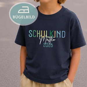 Könnte beinhalten: Marineblaues Kurzarm-T-Shirt mit dem Text "SCHULKIND Matteo 2026" in bunten Buchstaben. Eine türkis-weiße Grafik befindet sich in der oberen linken Ecke. Das T-Shirt wird von einem Kind getragen.