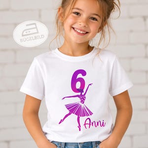 Könnte beinhalten: Ein weißes Kurzarm-T-Shirt mit einer Grafik aus einer violett-rosa verlaufenden Zahl 6, die in eine Ballerina-Silhouette im Tutu integriert ist, begleitet von dem rosa Schriftzug Anni, der unter der Tänzerin auf der Vorderseite gedruckt ist.