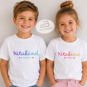 Könnte beinhalten: Zwei weiße T-Shirts mit kurzen Ärmeln und dem Wort Kitakind in einer Farbverlaufsschrift. Das linke Shirt zeigt den Namen Bennett mit Sternen, das rechte den Namen Emelie mit Herzen. Beide Shirts tragen ein kleines Bügeleisen-Logo.