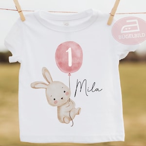 Könnte beinhalten: Ein weißes T-Shirt mit einer Grafik eines hellbraunen Hasen, der eine Schnur hält, die an einem rosa Luftballon mit der weißen Zahl 1 befestigt ist. Der Name Mila steht in schwarzer Schreibschrift neben dem Hasen. Das Shirt hängt an einer Leine.