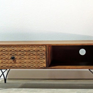 May include: A low-profile wooden media console featuring a single drawer with a textured geometric diamond pattern and a black knob. The unit includes an open storage compartment with a circular cable cutout and rests on four black metal legs.