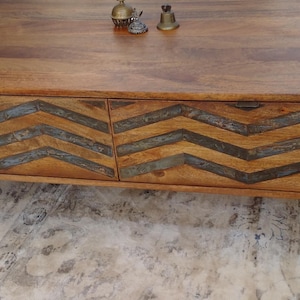 May include: A wooden coffee table with a natural finish. The front of the table features two drawers with a chevron pattern in a darker wood or metal. The table has metal legs and small decorative items on top.