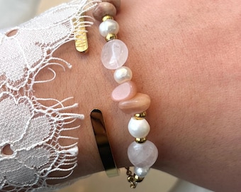 May include: A gold-toned metal cuff bracelet and a beaded bracelet featuring a mix of white pearls, translucent pink quartz stones, and smooth tan-coloured beads, all accented with small gold-toned spherical spacers arranged in a linear pattern.