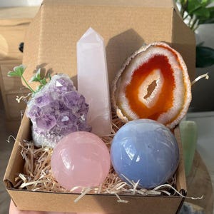 Può includere: Un set di cinque cristalli in una scatola di cartone con carta triturata. I cristalli includono una sfera di quarzo rosa, una sfera di celestite blu, un ammasso di ametista viola, una punta di quarzo rosa e una fetta di agata rossa.