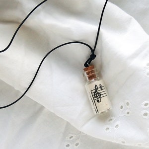 Puede incluir: Un collar con un cord&oacute;n negro y un peque&ntilde;o colgante de botella de vidrio. La botella contiene un trozo de papel con una clave de sol y notas musicales impresas en &eacute;l.