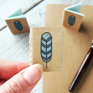 Puede incluir: Un pequeño cuaderno artesanal marrón con un diseño de pluma azul y blanca en la portada. El cuaderno está cerrado y está siendo sostenido por una mano.