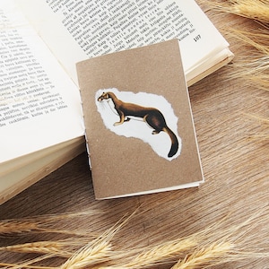 May include: A small brown notebook with a white paper cutout of a brown weasel on the front cover. The notebook is sitting on a wooden surface with wheat stalks on either side.