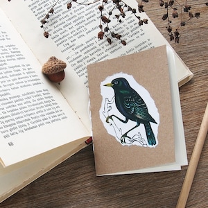 May include: A small, brown paper card with a black bird illustration. The bird is perched on a branch and has a dark green and blue color scheme. The card is sitting on a wooden surface with a book and a pencil.
