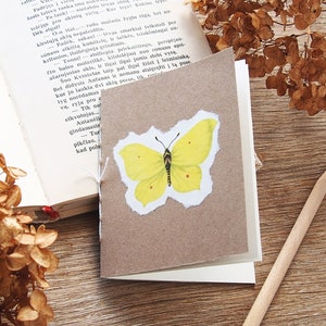 May include: A small, brown, handmade notebook with a yellow butterfly illustration on the front cover. The notebook is sitting on a wooden surface with a pencil and dried flowers.
