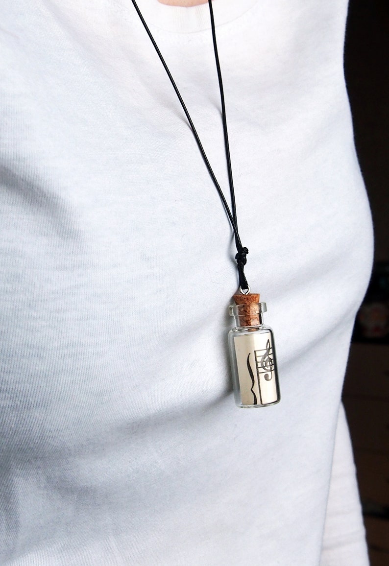 Puede incluir: Un collar con un cord&oacute;n negro y un peque&ntilde;o colgante en forma de botella de vidrio. La botella contiene un trozo de papel con una clave de sol y notas musicales impresas en &eacute;l.