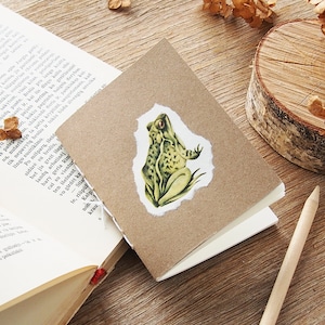 May include: A small, brown notebook with a green frog illustration on the front cover. The notebook is closed and sitting on a wooden surface.