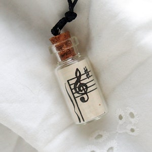 Puede incluir: Una peque&ntilde;a botella de vidrio con un tap&oacute;n de corcho, llena de un trozo de papel con una clave de sol y un pentagrama impresos. La botella est&aacute; unida a un cord&oacute;n negro, lo que la convierte en un collar.