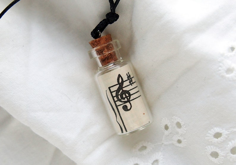 Puede incluir: Una peque&ntilde;a botella de vidrio con un tap&oacute;n de corcho, llena de un trozo de papel con una clave de sol y un pentagrama impresos. La botella est&aacute; unida a un cord&oacute;n negro, lo que la convierte en un collar.