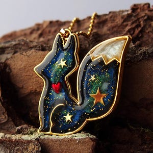 Puede incluir: Un collar de oro con un colgante con forma de zorro. El zorro está hecho de resina y tiene un diseño de galaxia azul y verde con estrellas doradas y un corazón rojo.
