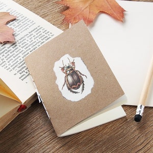 May include: A small, brown notebook with a beetle illustration on a torn paper label, resting on a wooden surface. An open book with aged pages and a pencil are also present, along with a dried leaf.